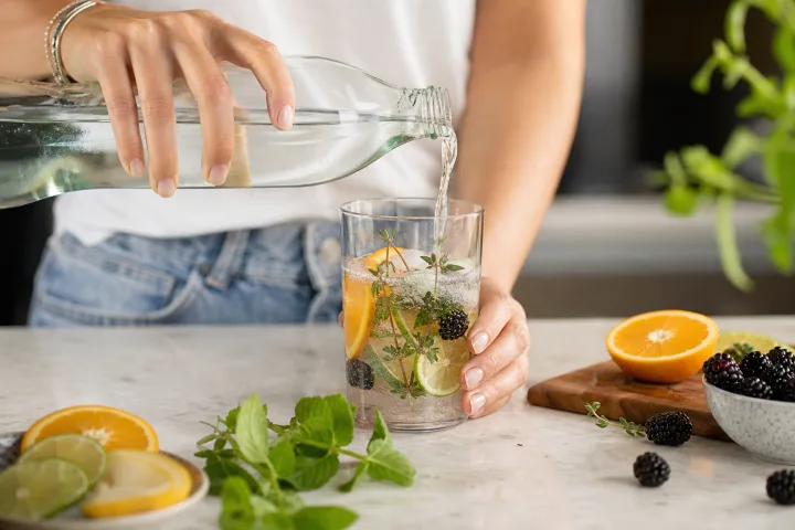 mani che versano acqua frizzante in un mocktail estivo con agrumi, more, erbe aromatiche e ghiaccio