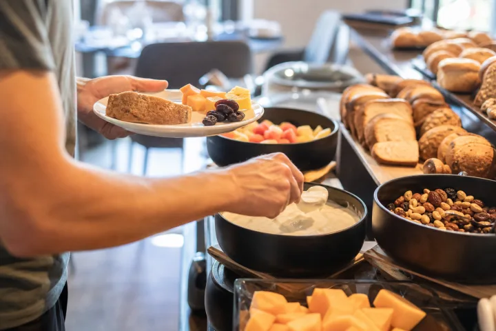 Mani al buffet dell'hotel compongono un piatto con yogurt, frutta e pane in una sala colazione luminosa
