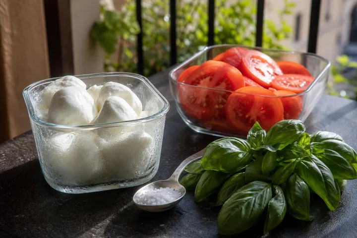 Contenitori separati con mozzarella fredda, pomodori e condimento su un tavolo estivo all’aperto