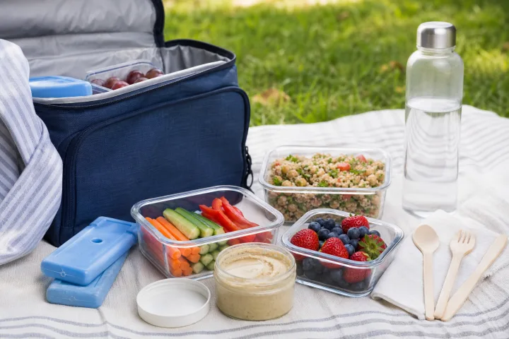 borsa frigo aperta con ghiaccioli gel, contenitori chiusi, frutta, verdure e borraccia su un telo pulito al parco.
