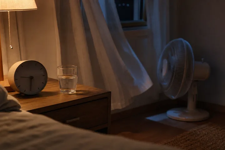 Comodino con bicchiere d'acqua e sveglia, tende leggere e ventilatore sullo sfondo rivolto verso la parete.