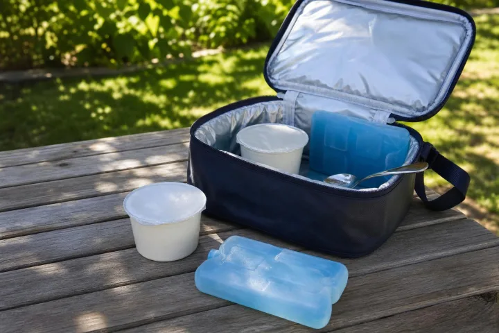 Borsa termica aperta su un tavolo estivo con yogurt sigillati, cucchiaio e ghiaccioli gel blu.