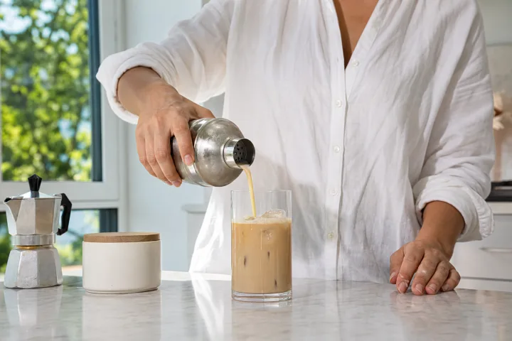 Persona che versa caffè shakerato in un bicchiere con ghiaccio in una cucina luminosa.