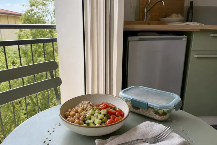 Ciotola estiva con cereali, ceci e verdure su un tavolino da balcone accanto a un contenitore riutilizzabile.