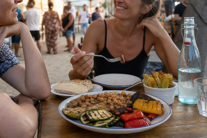 Tavolo all'aperto con verdure grigliate, legumi, pane, acqua e un piccolo assaggio fritto durante una sagra estiva.