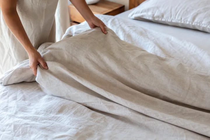 Mani che sistemano lenzuola chiare di cotone e lino su un letto estivo luminoso