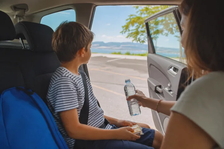 Bambino seduto nel sedile posteriore durante una pausa in auto con porta aperta e genitore che offre acqua.
