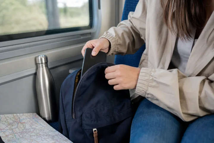 Mani che ripongono uno smartphone in uno zaino accanto a una borraccia e a una mappa su un sedile del treno.