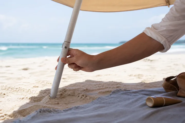Mano che regola l'inclinazione di un ombrellone su una spiaggia luminosa con telo e crema solare nella sabbia.