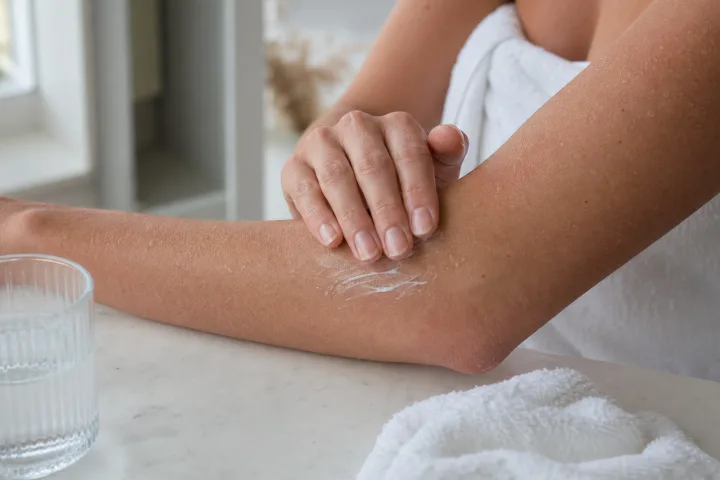 Mani che applicano crema idratante su un avambraccio asciutto accanto a un asciugamano chiaro.