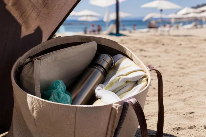 Borsa mare aperta sulla sabbia con sacca asciutta, borraccia, telo piegato e piccoli oggetti separati.
