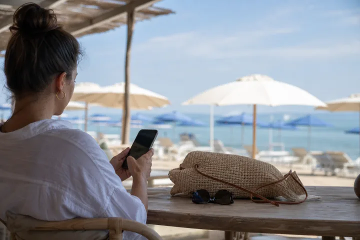Persona che usa uno smartphone su una terrazza balneare con ombrelloni sullo sfondo.